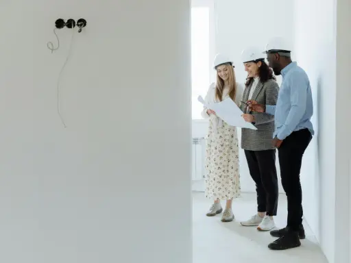 Tres arquitectos con cascos revisan planos en una obra, buscando cómo abaratar costes.