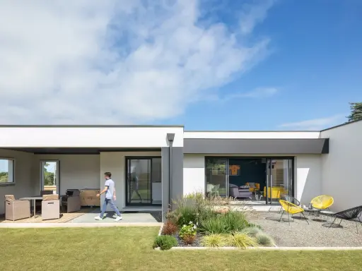 Une jeune personne traverse la pelouse devant une petite maison moderne avec une terrasse aménagée et un jardin de gravier.
