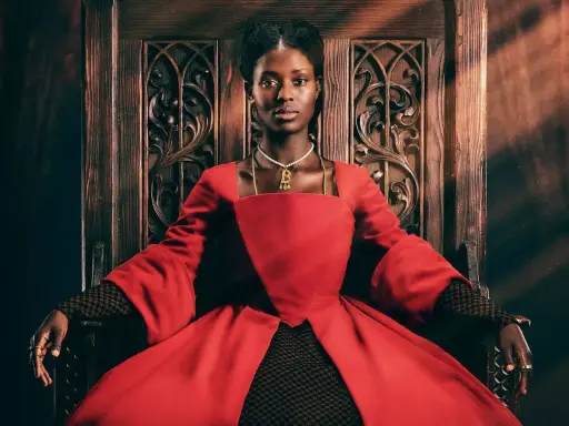 Jodie Turner-Smith, con su mirada regia, se sienta en un trono de madera tallada, vestida con un opulento traje rojo.