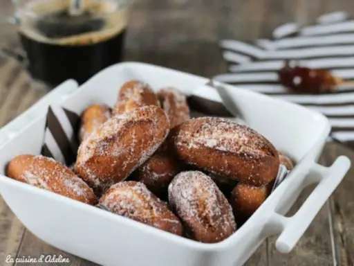 Beignets alsaciens schenkele dorés et saupoudrés de sucre, servis dans un plat blanc avec une tasse de café en arrière-plan.