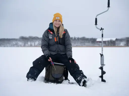 Kvinna ler och njuter av att fiska på isen. Bredvid henne står en isborr redo för användning.
