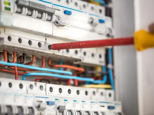 Ręka z czerwonym śrubokrętem pracuje przy instalacji niskoprądowej. Widać bezpieczniki i kolorowe przewody.