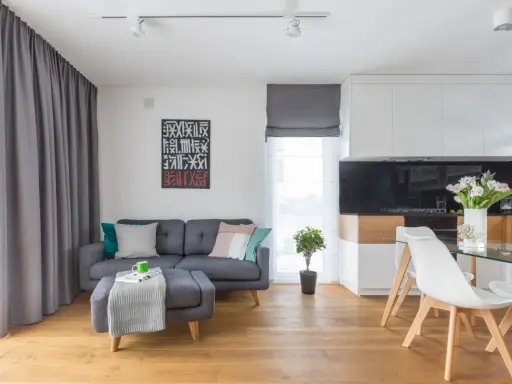 Mały salon inspiracje: szara kanapa, stolik z pufą, nowoczesna kuchnia i jadalnia. Drewniana podłoga dodaje ciepła.