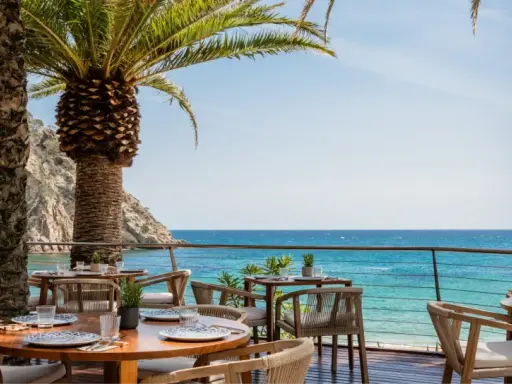 Mesas de restaurante con vistas al mar en el pola giverola resort, bajo palmeras.
