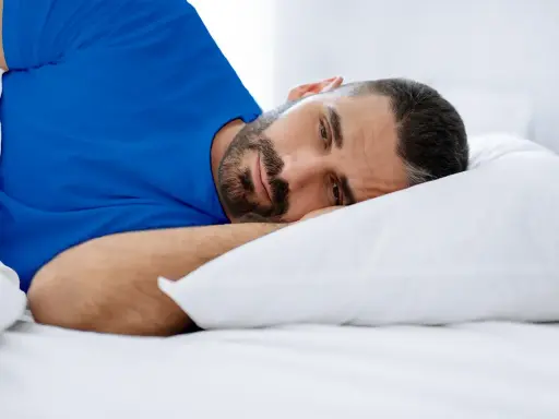 Hombre con barba en cama, con ojos abiertos, vistiendo camiseta azul. Duermo pero no descanso, ¿qué puedo hacer?