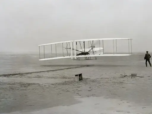 Der Wright Flyer hebt vom sandigen Boden ab, ein Mann beobachtet den historischen Flug.
