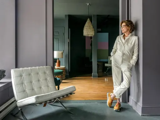 A woman in a cream jumpsuit leans against a wall in the robin rice gallery, with a Barcelona chair and abstract art visible.