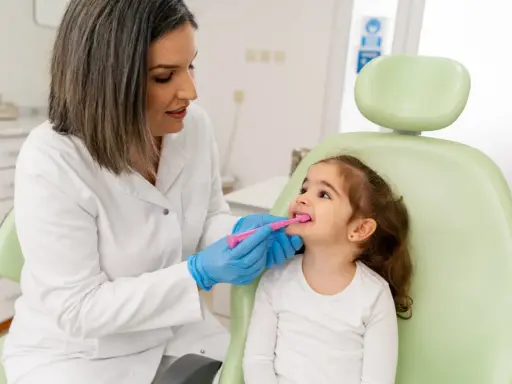 Dentystka w białym fartuchu i niebieskich rękawiczkach pokazuje małej dziewczynce, jak prawidłowo szczotkować zęby. To przykład troski o zdrowie jamy ustnej w ramach pedodoncji.