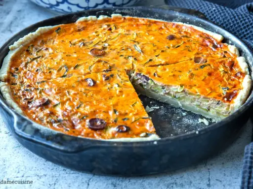 Herzhaftes Quiche Rezept mit Zucchini und Oliven, goldbraun gebacken in einer Gusseisenpfanne. Ein Stück ist bereits herausgeschnitten.