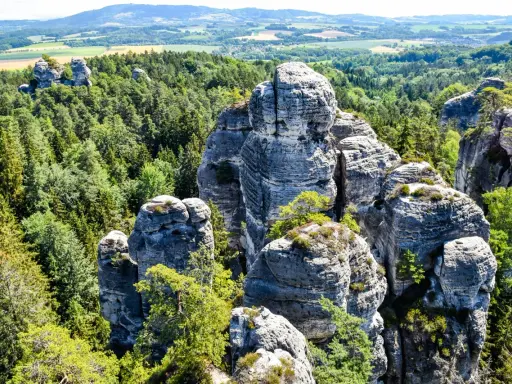 Majestatyczna, hruba skala wznosi się pośród gęstego lasu. W oddali widać pagórkowaty krajobraz.