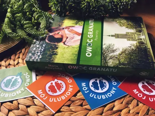 Książka "Owoc granatu" Marii Paszyńskiej z motywem pary na okładce, otoczona zielenią i kolorowymi naklejkami z symbolem granatu.