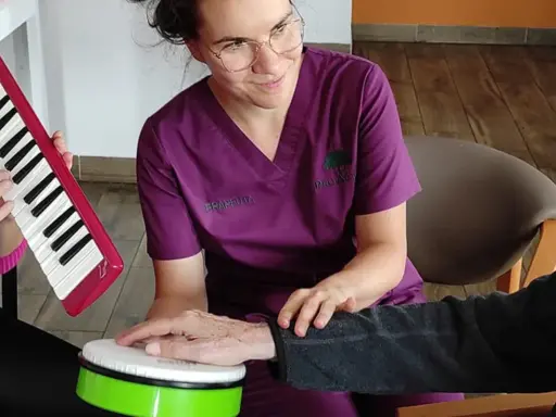 Terapia dźwiękiem i rytmem: muzykoterapia dla seniorów. Terapeuta wspiera dłoń pacjenta na bębenku, podczas gdy inny uczestnik gra na keyboardzie.