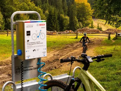Stacje ładowania rowerów elektrycznych w Piwnicznej-Zdroju. W tle rowerzystka na szlaku.