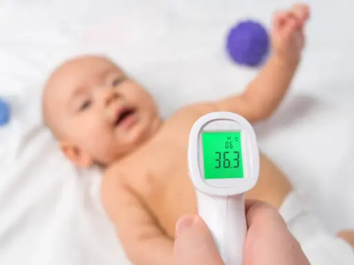 Jak mierzyć temperaturę termometrem elektronicznym? Dziecko leży na białym tle, a termometr pokazuje 36.3°C.