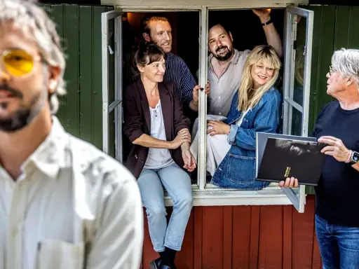 En man med gula glasögon och en grupp människor i ett fönster. En man håller en bok med texten "Allan Edwall barn".