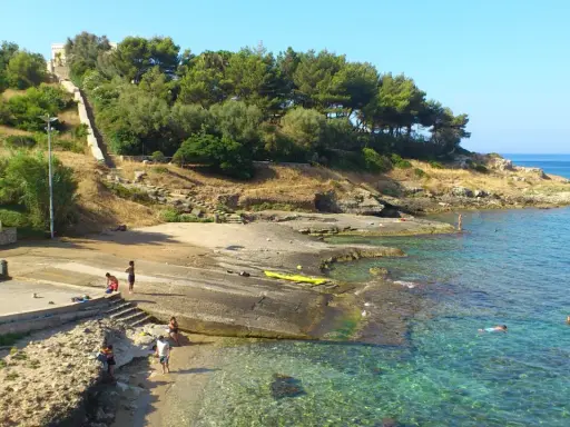 Scogliera e mare cristallino a San Gregorio Leuca. Persone si godono il sole e l'acqua, una scalinata sale verso la collina alberata.