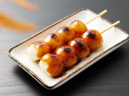 Brochettes de dango grillés, nappées de sauce sucrée, sur une assiette blanche.