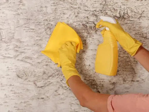 Manos con guantes amarillos limpiando paredes con un paño y spray.