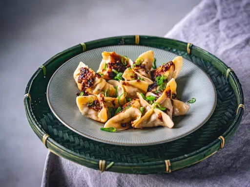 Gyoza vegetarisch, appetitlich mit würziger Soße und Frühlingszwiebeln garniert, auf einem Teller in einem grünen Bambuskorb serviert.