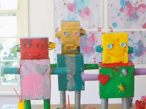 Drei bunte Roboter, die Kinder gebastelt haben, stehen nebeneinander. Sie sind aus Kartons und Rollen gefertigt und mit leuchtenden Farben bemalt.