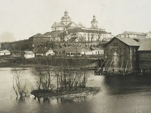 Widok na miasto z rzeką i kościołem. W oddali widać potężną budowlę sakralną z kopułami. Na pierwszym planie rzeka z wysepką i drzewami. Po prawej stronie drewniane budynki nad wodą. Ktoś pisze na Berdyczów.