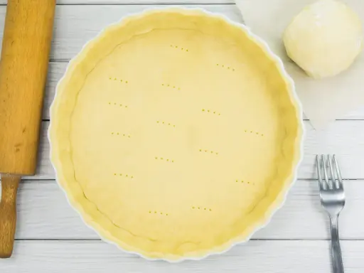 Une pâte brisée est prête dans une casserole blanche, avec une boule de pâte, une rouleau et une fourchette à côté.