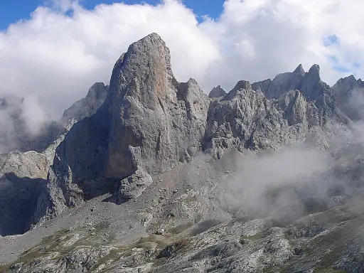Majestatyczny **Naranjo de Bulnes** w Picos de Europa, otoczony mgłą i chmurami, z małym schroniskiem u podnóża.
