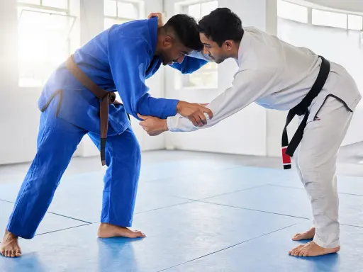 Dwóch mężczyzn w kimonach do BJJ, jeden w niebieskim z brązowym pasem, drugi w białym z czarnym pasem, przygotowuje się do walki.