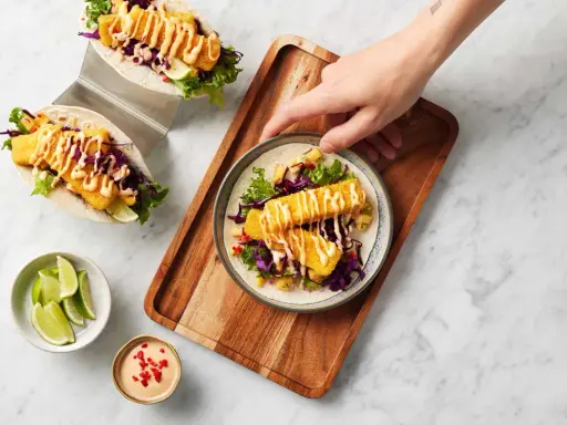 Två läckra fish tacos med krispig fisk, rödkål, sallad och en krämig sås. En hand sträcker sig efter en tallrik med en taco.