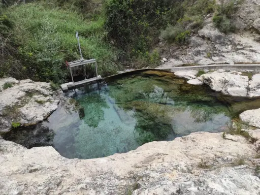 Aguas cristalinas en los baños de Somogil, rodeadas de rocas y vegetación. Una compuerta metálica controla el flujo del agua.