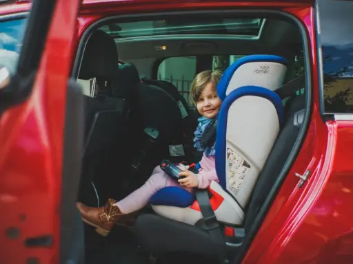 Uśmiechnięta dziewczynka w foteliku samochodowym Britax Römer w czerwonym aucie.