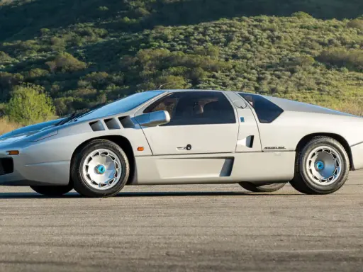 Silberner Isdera Imperator 108i mit markanten Lufteinlässen und futuristischem Design, geparkt vor einer grünen Hügellandschaft.
