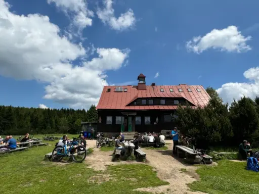 Chatka górzystów z czerwonym dachem, otoczona lasem. Ludzie odpoczywają przy stołach, obok stoją rowery.