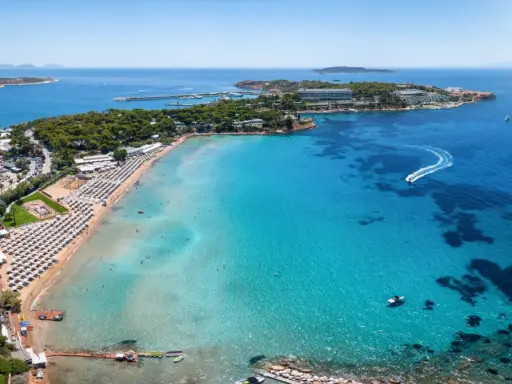 Traumhafter Blick auf das athen meer mit einer belebten Strandpromenade, Liegestühlen und dem glitzernden, türkisfarbenen Wasser, auf dem Boote fahren.