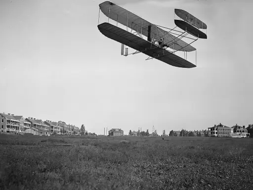 Das Flugzeug der Gebrüder Wright fliegt über ein Feld. Im Hintergrund sind Gebäude zu sehen.