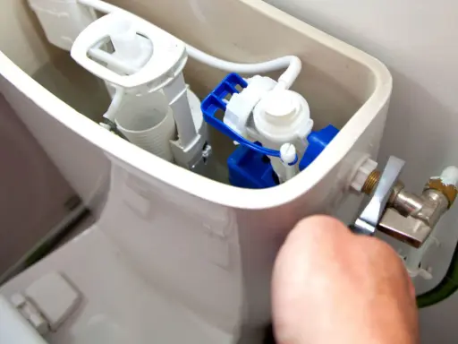 Mécanisme de chasse d'eau qui coule, avec un flotteur bleu et blanc visible. Une main ajuste le robinet d'arrivée d'eau.