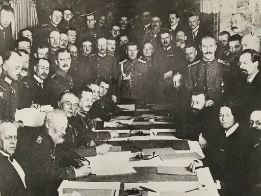 Delegierte unterzeichnen den Frieden von Brest-Litowsk. Männer in Uniformen und Anzügen sitzen an einem Tisch, umgeben von weiteren Personen.