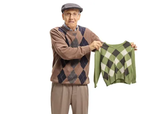 Äldre man med argyle-tröja håller upp en mindre, likadan tröja. Han ser bekymrad ut, som om bomullen krymper.