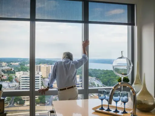 Hombre mira por ventana de aluminio, pensando cuánto pesa una ventana de aluminio. Vista urbana y río.