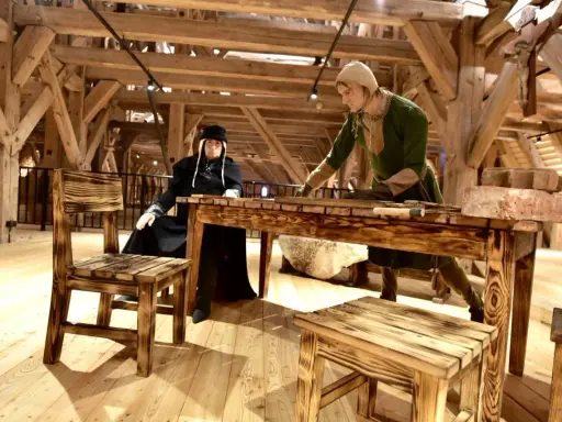 W muzeum Mazury dwie figury w strojach historycznych przy drewnianym stole. Jedna siedzi, druga stoi, pracując.
