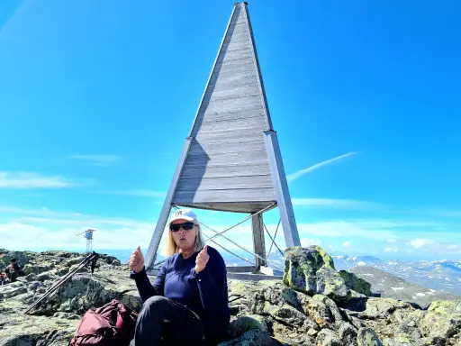 En kvinna sitter på toppen efter att ha lyckats bestiga Helags. Hon ger tummen upp vid den ikoniska träpyramiden.