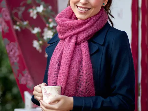 En kvinna ler och håller en mugg, insvept i en mjuk, rosa mohairhalsduk.