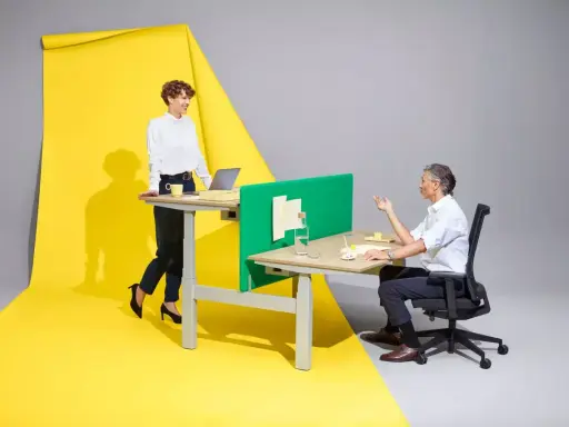 Femme en position debout statique, souriante, devant un bureau. Homme assis à un bureau, discutant. Fond jaune et gris.