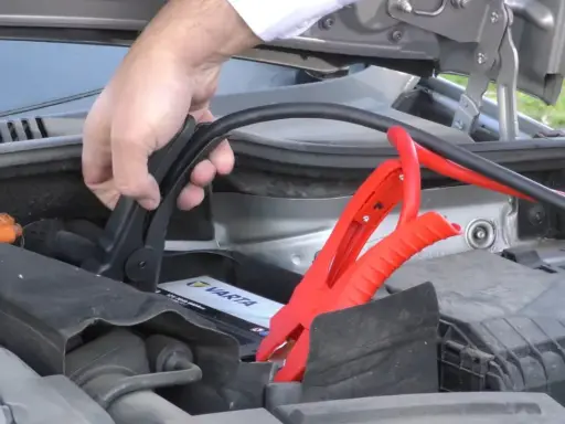 Mano conectando pinzas rojas a la batería de un coche para arrancar con pinzas.