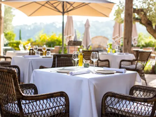 Tables dressées en terrasse, sous un parasol, invitant à savourer une cuisine du terroir authentique. Vins, verres et couverts attendent les convives.