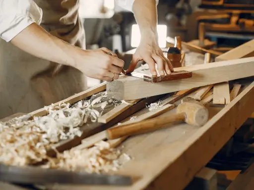 Un artisan qui travaille le bois, concentré sur la précision de ses gestes. Des copeaux de bois jonchent l'établi.
