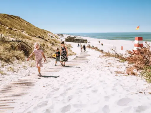 Familj på väg längs en träspång över vit sand i Danmark. En rödvit fyr och bunkrar syns i bakgrunden.