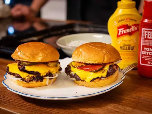 Två saftiga hamburgare med smält ost, pickles och lök på en tallrik. Bredvid står flaskor med senap och ketchup.