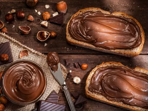 Selbstgemachte Haselnusscreme auf Brot, umgeben von Nüssen und Schokolade. Ein Genuss, der zum Nachmachen einlädt.