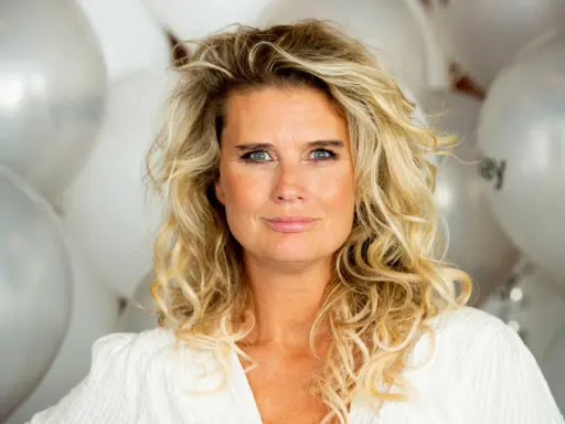 Blond kvinna med blå ögon och lockigt hår, omgiven av silverfärgade ballonger. Hon ser ut att vara redo för festliga udda veckor.